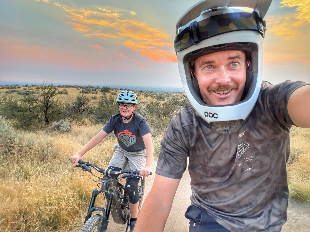 Jed in a full-face helmet with a young mountain biker riding behind him