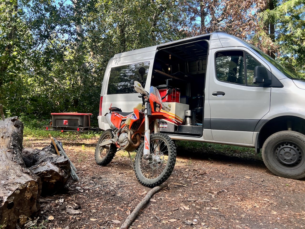 An orange dirt bike in front of the open van at a wooded campsite