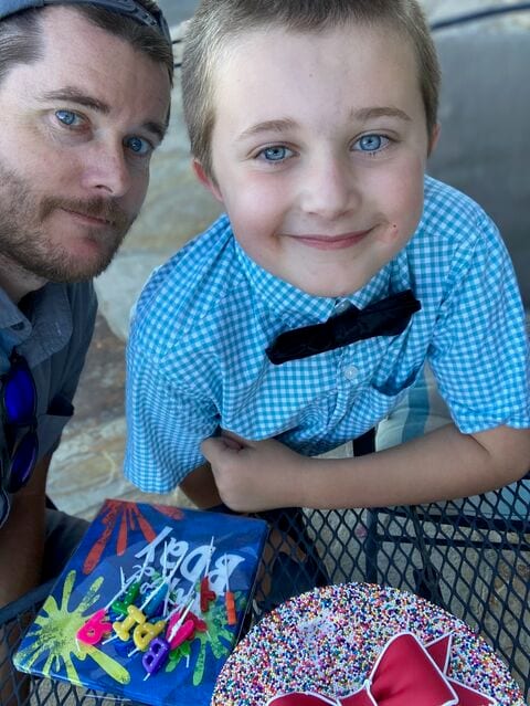 Jed and his young son in a blue checked shirt and bow tie, with a birthday cake covered in sprinkles and a pile of letter candles