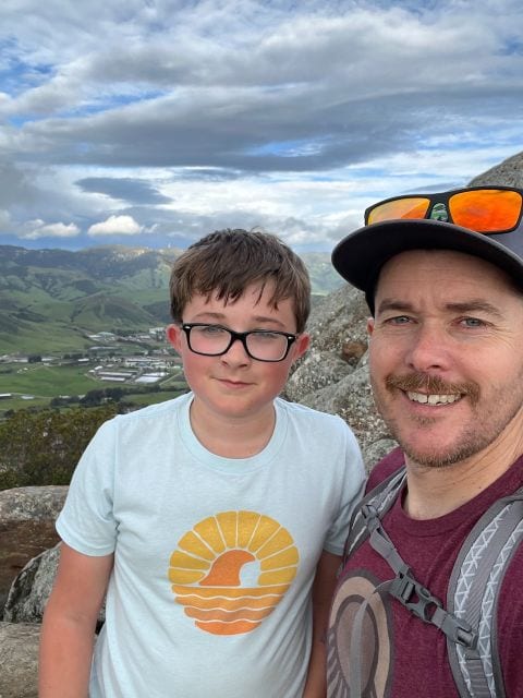 Jed and his son, now older and wearing glasses, in a selfie on a rocky hilltop above green valleys